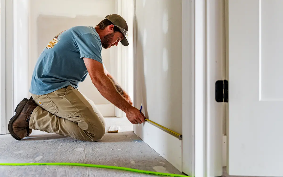 Builder kneeling to measure door trim with a tape measure