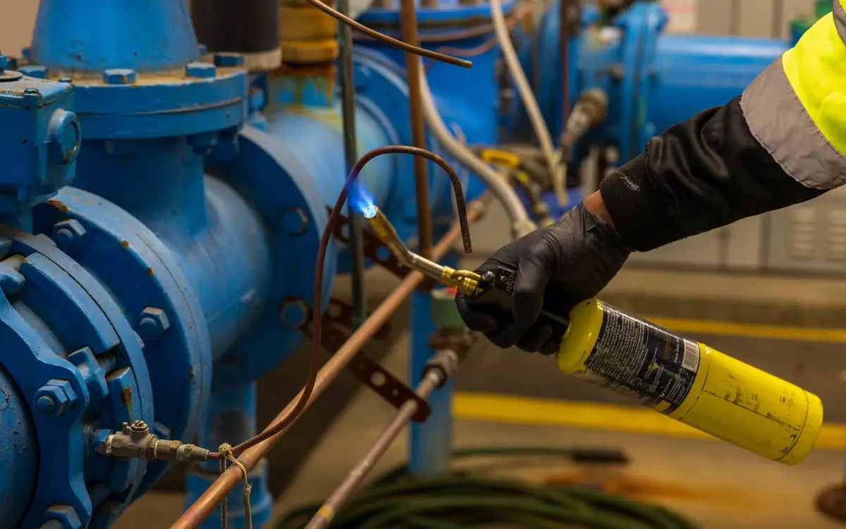 Gloved hand using a gas torch on a copper pipe beside blue pipework