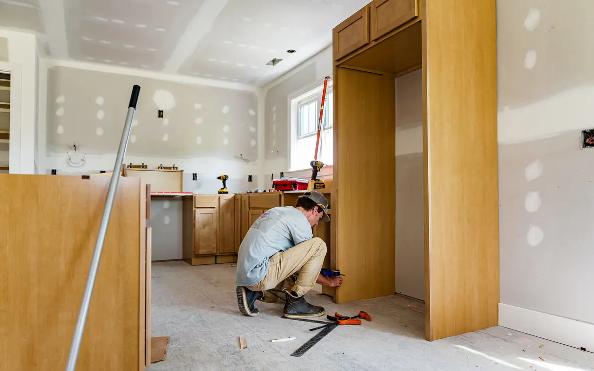 Carpenter fitting kitchen cabinets in a room with freshly taped drywall